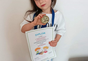Dziewczynka stoi z dyplomem i medalem.